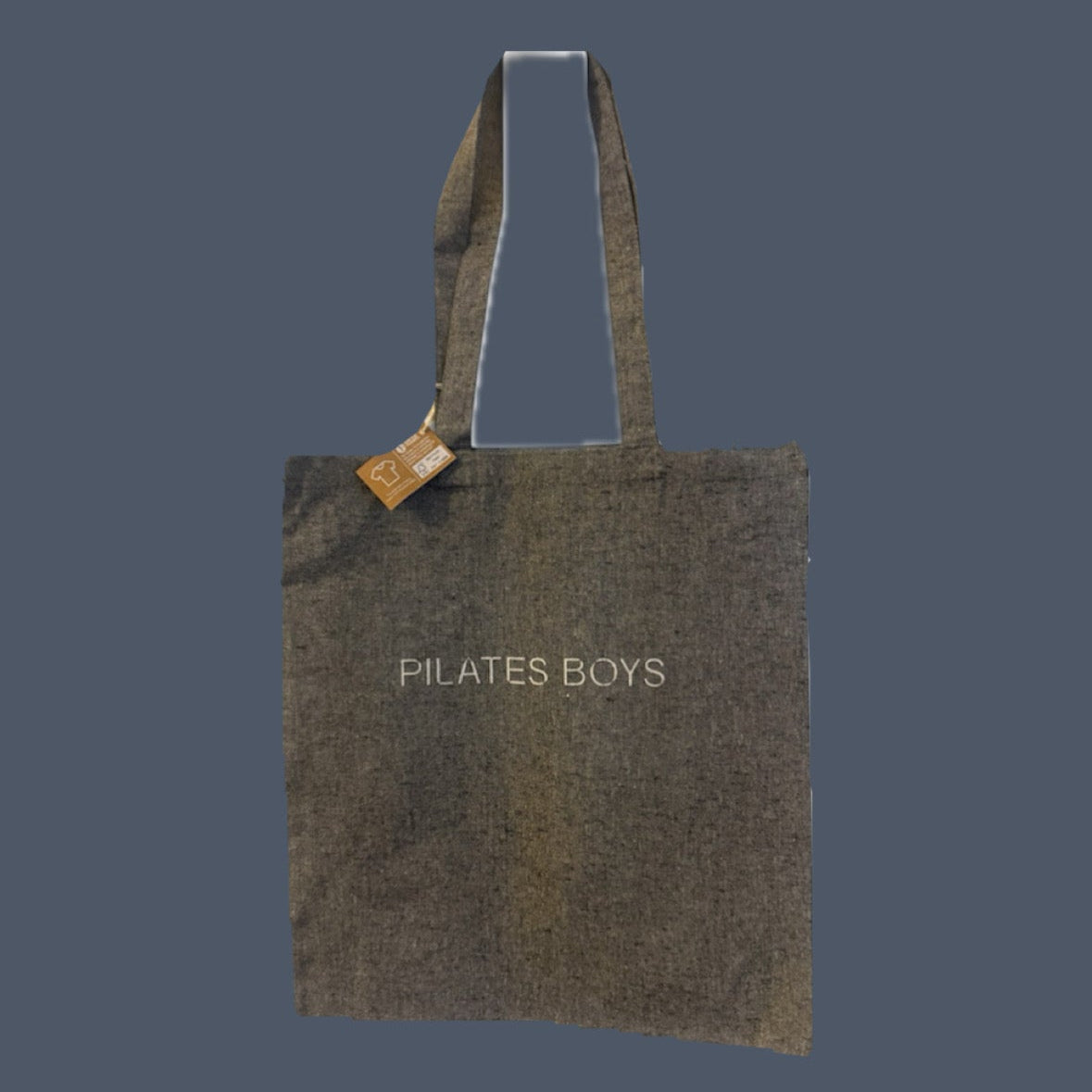 A grey canvas tote bag with 'PILATES BOYS' printed in white text on the front, featuring dual top handles and a small wooden brand tag, set against a solid dark slate blue background.