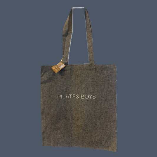 A grey canvas tote bag with 'PILATES BOYS' printed in white text on the front, featuring dual top handles and a small wooden brand tag, set against a solid dark slate blue background.